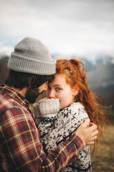 Side view of tender couple hugging and bonding with man closed eyes and woman looking at camera in cold day in mountains - ADSF12043