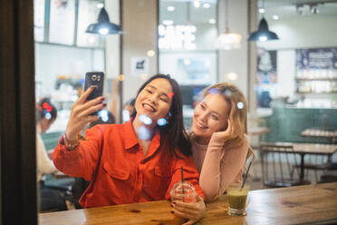 Zwei junge Frau Freunde mit Tassen frischen Kaffee lächelnd und posieren für selfie beim Sitzen am Tisch in gemütlichen Restaurant mit einem Getränk - ADSF12046