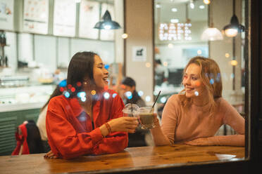 Junge gemischtrassige Frauen, die lächelnd miteinander sprechen, während sie an einem Tisch in einem gemütlichen Café sitzen und mit Tassen anstoßen und Fruchtgetränke genießen - ADSF12047