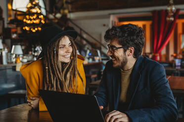 Kluger Mann mit Brille und stilvolle Frau mit Hut schauen auf einen Monitor, während der Mann in einem Café auf einem Laptop tippt - ADSF12050