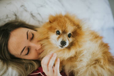 Junge glückliche lächelnde attraktive Frau im Pyjama im Bett liegend mit kleinem flauschigen Hund - ADSF12063