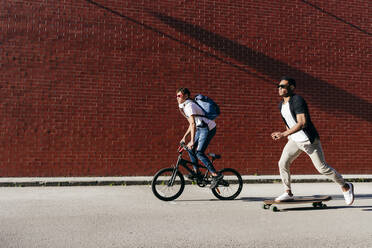 Seitenansicht junger schwarzer erfolgreicher männlicher Freunde in lässiger Kleidung und mit stilvoller Sonnenbrille, die Fahrrad und Skateboard fahren - ADSF12101