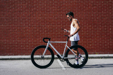 Seitenansicht eines gut aussehenden männlichen Radfahrers in Sportkleidung und mit Sonnenbrille, der sein Smartphone benutzt, während er mit seinem Fahrrad neben einer roten Backsteinmauer steht - ADSF12111