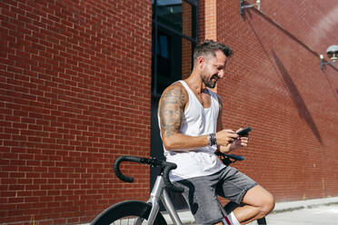Gut aussehender männlicher Radfahrer in Sportkleidung, der sein Smartphone benutzt, während er sich auf dem Fahrrad neben einer roten Backsteinmauer ausruht - ADSF12112