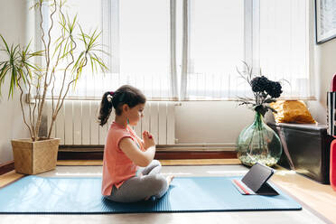 Kleines Mädchen sitzt im Lotussitz und meditiert auf der Matte mit geschlossenen Augen zu Hause mit großem Fenster, während sie Yoga übt und Video-Tutorials auf dem Tablet sieht - ADSF12114