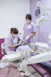 Female dentist in uniform and mask curing patient teeth with female assistant in modern dental clinic - ADSF12120