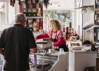 Ein älterer Mann wählt Lebensmittel aus der Tiefkühltruhe aus, während er Wein und Käse von einer Verkäuferin in einem gemütlichen kleinen Delikatessenladen kauft - ADSF12127