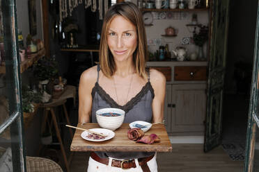 Lächelnde schöne Frau hält Tablett mit Matcha-Tee und Trockenblumen im Café - RDGF00101