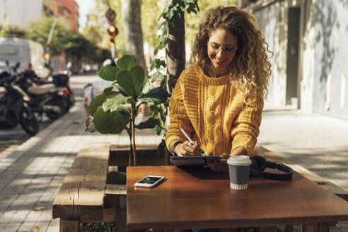 Smiling beautiful female student using digital tablet while sitting at sidewalk cafe in city - BOYF01345