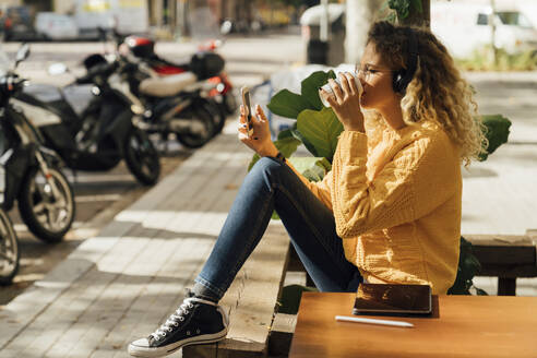 Young female student drinking coffee while using smart phone at sidewalk cafe in city - BOYF01349