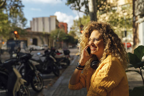 Glückliche junge Studentin mit lockigem blondem Haar, die mit einem Smartphone in der Stadt spricht - BOYF01354