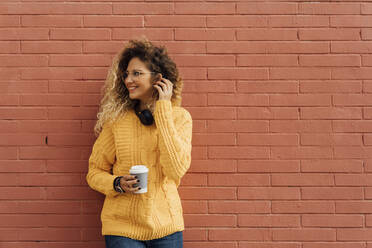 Smiling female student with long curly hair looking away while holding disposable coffee cup against red brick wall - BOYF01360