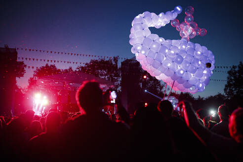 Wal aus lilafarbenen Luftballons schwebt über einer Menschenmenge, die sich bei einem nächtlichen Musikfestival amüsiert - SAJF00067