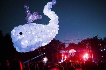 Wal aus lilafarbenen Luftballons schwebt über einer Menschenmenge, die sich bei einem nächtlichen Musikfestival amüsiert - SAJF00068