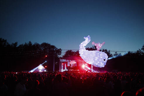 Wal aus lilafarbenen Luftballons schwebt über einer Menschenmenge, die sich bei einem nächtlichen Musikfestival amüsiert - SAJF00069