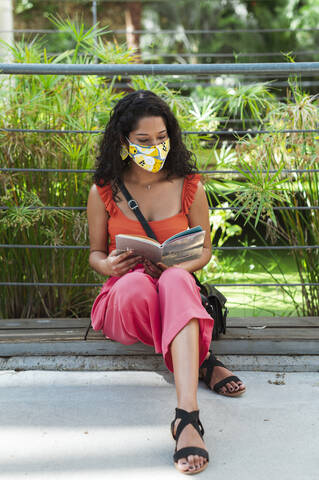 Young woman wearing protective mask while reading book sitting in park during COVID-19 pandemic stock photo