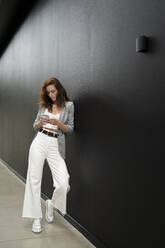 Geschäftsfrau lehnt sich an eine schwarze Wand und benutzt ein Smartphone im Korridor - MTBF00601