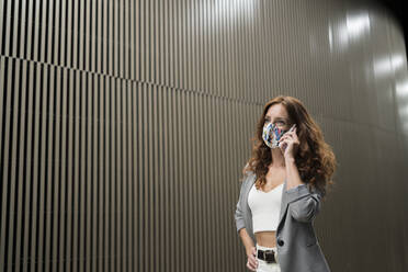 Female professional wearing protective mask while talking on smart phone against striped wall in corridor - MTBF00607