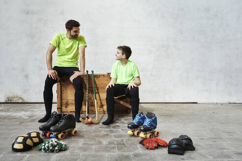 Father and son talking while sitting on wooden box by sports equipment against wall at court - VEGF02811