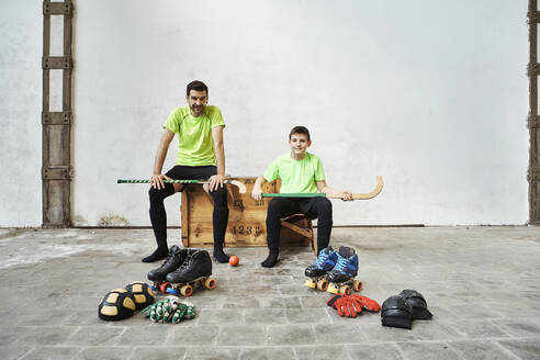 Father and son sitting with hockey sticks on wooden box by sports equipment against wall at court - VEGF02813