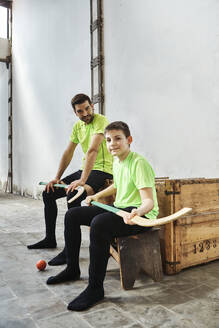 Confident boy and father sitting with hockey sticks against wall at court - VEGF02814