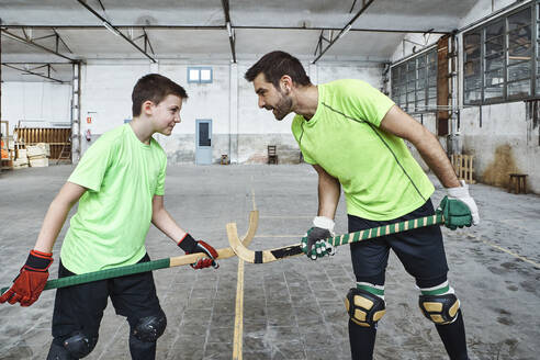 Father and son holding hockey sticks while standing face to face while playing on court - VEGF02830