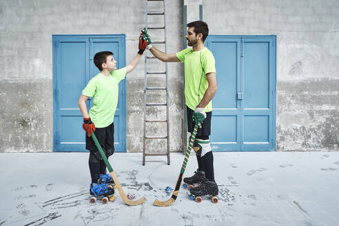 Father and son holding hockey sticks while giving high-fives against doors at court - VEGF02837