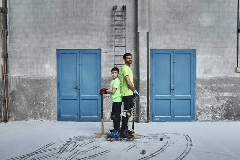 Father and son standing back to back while holding hockey sticks by doors at court - VEGF02838