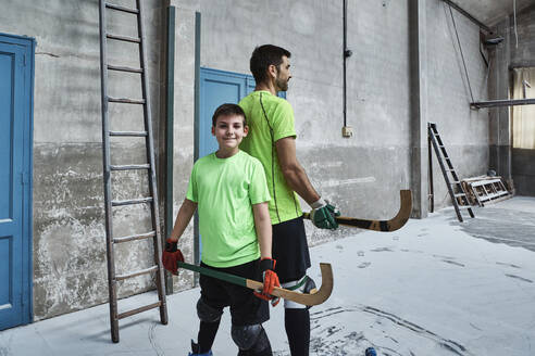 Smiling boy holding hockey stick while standing back to back with father at court - VEGF02839