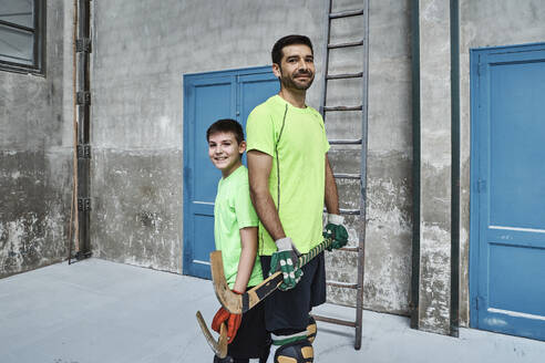 Smiling boy and father holding hockey sticks while standing back to back at court - VEGF02840