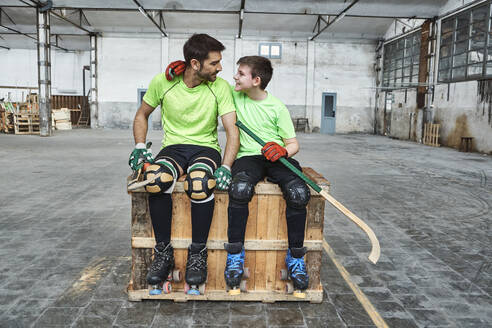 Smiling boy with arm around father while sitting with hockey sticks on wooden box at court - VEGF02848