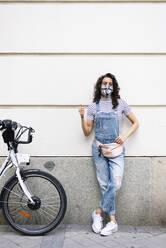 Mature woman wearing protective face mask showing thumbs up while standing by electric bicycle against wall - JCMF01206