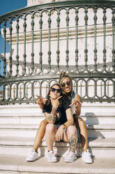 Female friends gesturing while sitting on staircase against fence at Praca Do Comercio, Lisbon, Portugal - DCRF00697