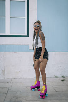 Cheerful woman with roller skates on footpath against wall in city - DCRF00700