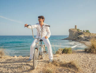 Älterer bärtiger sportlicher Mann mit Sonnenbrille beim Radfahren am Meer mit trockenem Gras auf dem Hintergrund der erstaunlichen türkisfarbenen Meereslandschaft am hellen Tag - ADSF12175