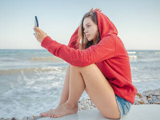 Young smiling woman with long hair in red hoodie taking selfie on smartphone lying on surfboard on stony seaside - ADSF12176