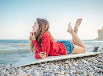 Seitenansicht einer attraktiven, lächelnden, langhaarigen Frau in rotem Kapuzenpulli, die es genießt, auf einem Surfbrett am steinigen Meeresufer zu liegen - ADSF12177
