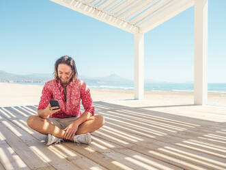 Bearded male browsing in smartphone while sitting in gazebo on sandy beach near sea on sunny day - ADSF12183