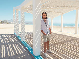 Handsome bearded man keeping hands in pockets and looking away while leaning on pillar of white gazebo on sandy beach - ADSF12189