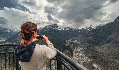 Back view woman in warm clothes standing on pier and looking at mountain ridge and taking picture with mobile phone in countryside - ADSF12191