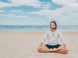 Sportlicher bärtiger Mann, der am ruhigen Meeresufer trainiert und Yoga-Asanas gegen das blaue Meer und den Himmel macht - ADSF12192