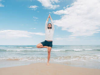 Sportlicher bärtiger Mann, der am ruhigen Meeresufer trainiert und Yoga-Asanas gegen das blaue Meer und den Himmel macht - ADSF12194