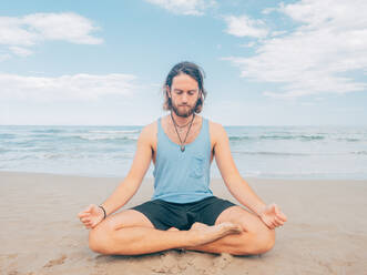 Sportive bearded male training on tranquil seashore and doing yoga asana against blue sea and sky - ADSF12195