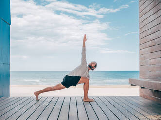 Erwachsener, fitter, bärtiger Mann macht Yoga auf einem Holzsteg am Meer und schaut weg - ADSF12198