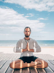 Adult bearded male meditating while sitting in lotus pose on wooden pier by seashore with legs crossed and looking at camera - ADSF12199