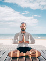 Erwachsener bärtiger Mann meditiert im Lotussitz auf einem Holzsteg am Meer mit gekreuzten Beinen und Blick in die Kamera - ADSF12199