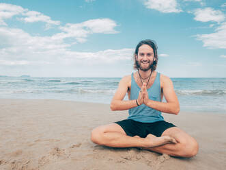Sportlicher bärtiger Mann, der am ruhigen Meeresufer trainiert und Yoga-Asanas gegen das blaue Meer und den Himmel macht - ADSF12200