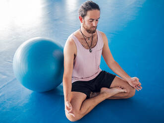 Bärtiger Mann in Sportkleidung mit geschlossenen Augen und gekreuzten Beinen meditiert auf blauem Boden mit Gymnastikbällen im Studio - ADSF12201
