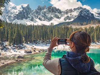 Rückenansicht der jungen Frau in Daunenweste Schießen auf Telefon majestätische Landschaft von türkisfarbenen Wasser durch felsige Berge und Wald in bewölkten Tag umgeben - ADSF12213