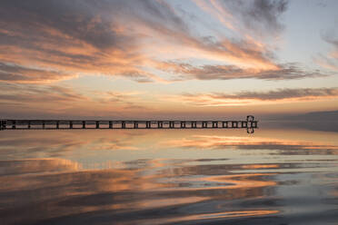 Silhouettes of distant unrecognizable people standing on pier near tranquil lake against sunset sky - ADSF12225